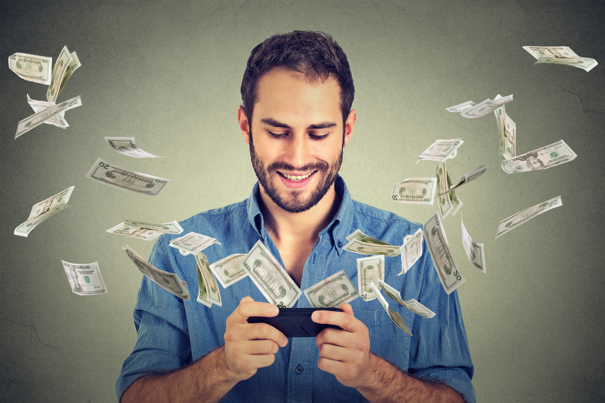 Image of guy wearing a blue shirt smiling while looking at his cell phone and money bills are flying around.